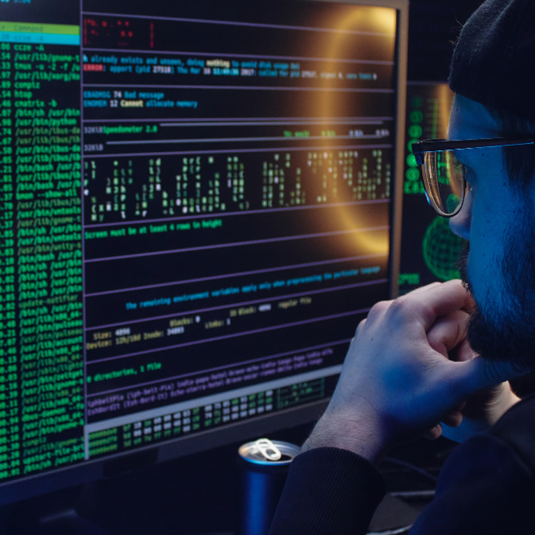 bearded man with cap and glasses looking at multiple monitors with dark screens and colorful numbers and characters 