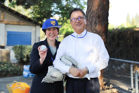 Two people smiling; one is wearing a hat and holding a baseball