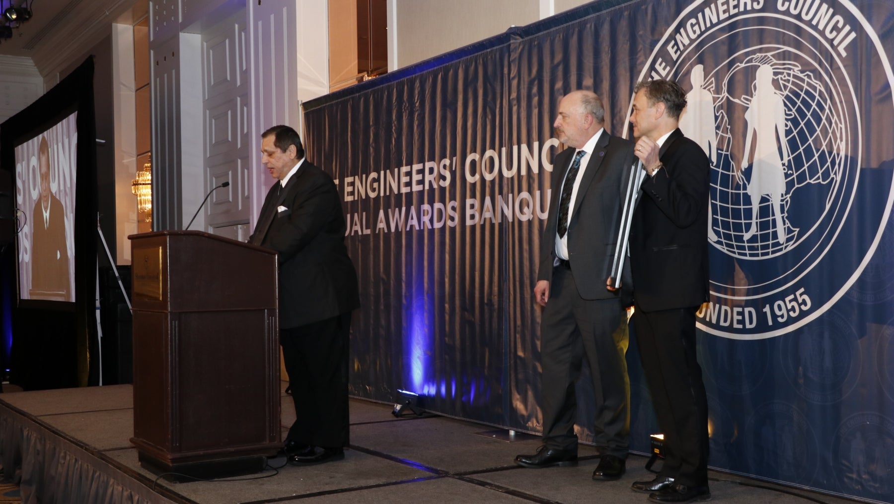 Ozkan stands at a podium next to two other people watching him give acceptance speech 