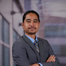 Male student wearing a grey suit with a blue shirt and striped tie with arms crossed looking straight into the camera. 