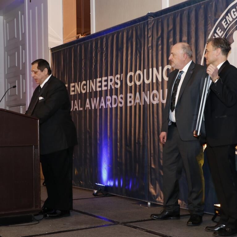 Ozkan stands at a podium next to two other people watching him give acceptance speech 