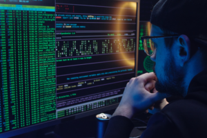 bearded man with cap and glasses looking at multiple monitors with dark screens and colorful numbers and characters 