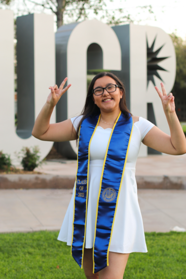 Woman wearing a graduation stole throws up the peace sign in front of the big UCR statue letters